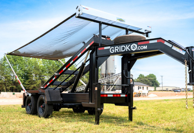 Black Gridko gooseneck roll-off trailer with its tarp cover system extended, parked on grass with an industrial facility and silos in the background.
